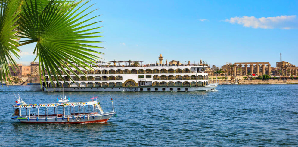 Cruise ship and small boat sailing on the Nile River in Luxor, Egypt, with ancient temple ruins and palm leaves in the background.