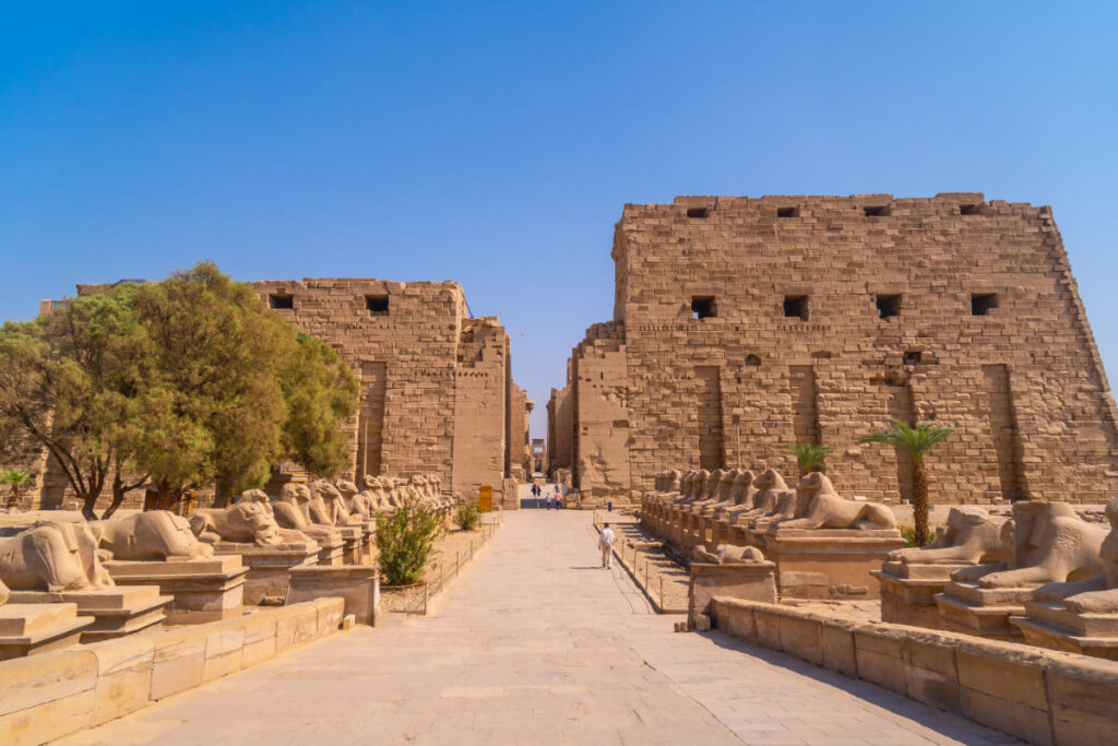 Entrance of Karnak Temple in Luxor, Egypt, with rows of ram-headed sphinx statues and massive sandstone pylons under a clear blue sky.