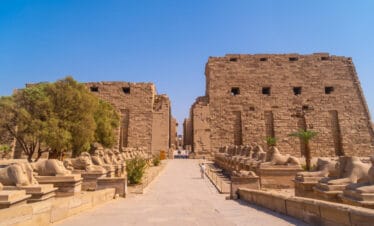 Entrance of Karnak Temple in Luxor, Egypt, with rows of ram-headed sphinx statues and massive sandstone pylons under a clear blue sky.