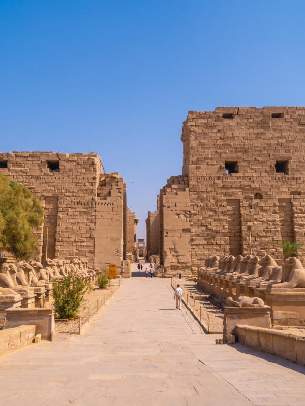 Entrance of Karnak Temple in Luxor, Egypt, with rows of ram-headed sphinx statues and massive sandstone pylons under a clear blue sky.