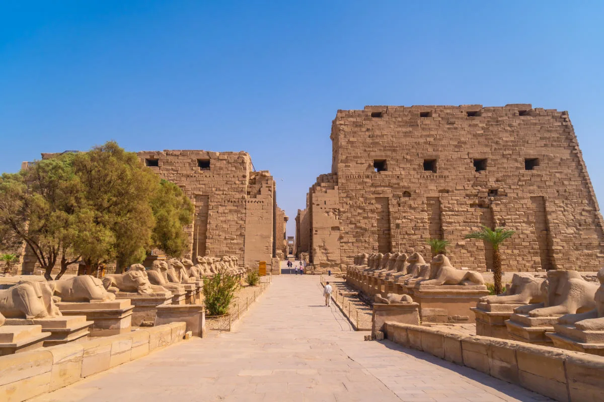 Entrance of Karnak Temple in Luxor, Egypt, with rows of ram-headed sphinx statues and massive sandstone pylons under a clear blue sky.