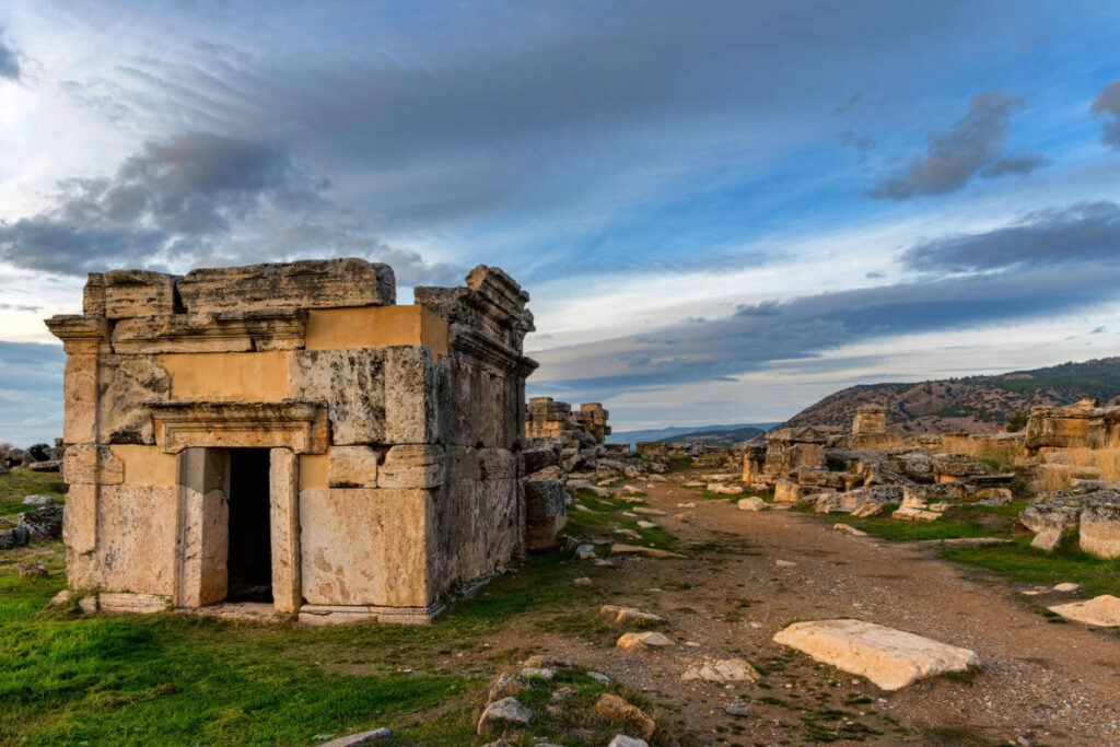 Ancient stone tomb structure in the necropolis of Hierapolis, Pamukkale, with dramatic skies and scattered ruins.