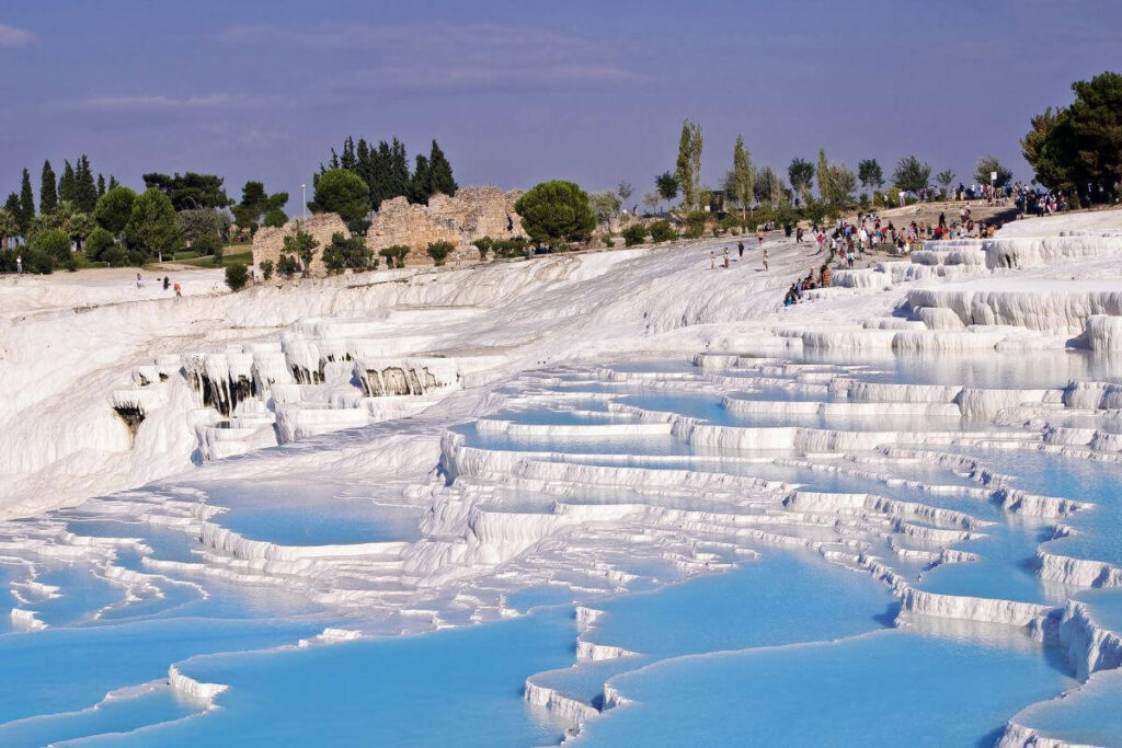 Blue thermal pools cascading down white travertine terraces in Pamukkale, Turkey, with tourists exploring the site.