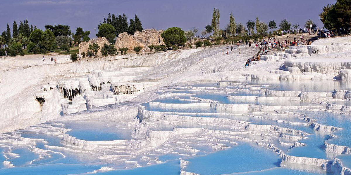 Blue thermal pools cascading down white travertine terraces in Pamukkale, Turkey, with tourists exploring the site.