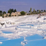 Blue thermal pools cascading down white travertine terraces in Pamukkale, Turkey, with tourists exploring the site.