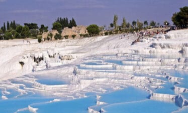 Blue thermal pools cascading down white travertine terraces in Pamukkale, Turkey, with tourists exploring the site.
