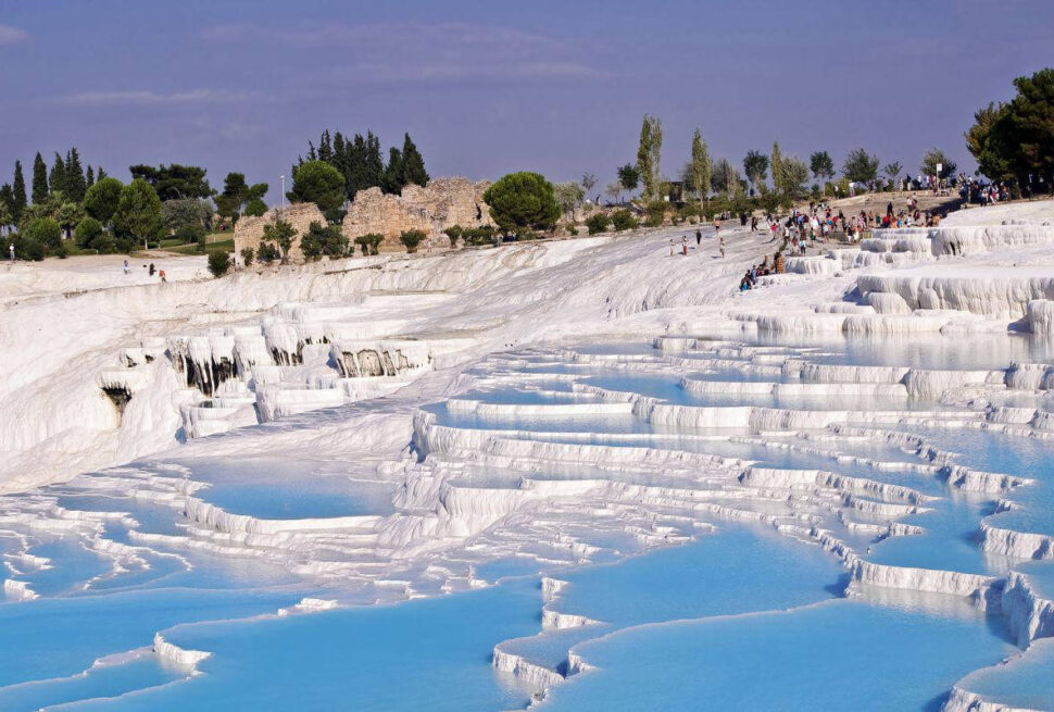 Blue thermal pools cascading down white travertine terraces in Pamukkale, Turkey, with tourists exploring the site.