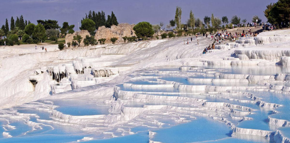 Blue thermal pools cascading down white travertine terraces in Pamukkale, Turkey, with tourists exploring the site.