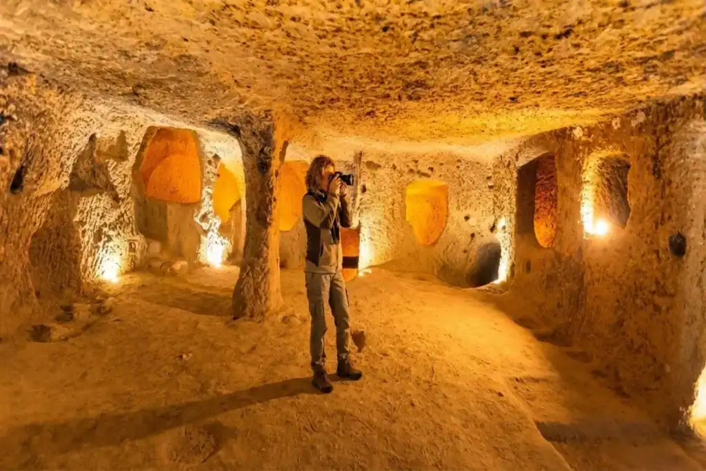Best Cappadocia Tours from Istanbul (Local Agency Guide – 2026) 2 A female tourist with a camera exploring the intricately carved, warmly lit chambers of an ancient underground city, such as Derinkuyu or Kaymakli, in Cappadocia, Turkey.
