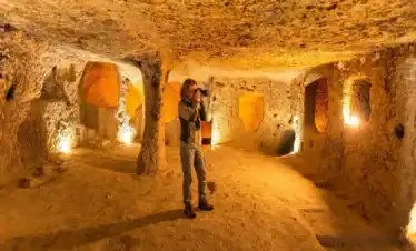 A female tourist with a camera exploring the intricately carved, warmly lit chambers of an ancient underground city, such as Derinkuyu or Kaymakli, in Cappadocia, Turkey.
