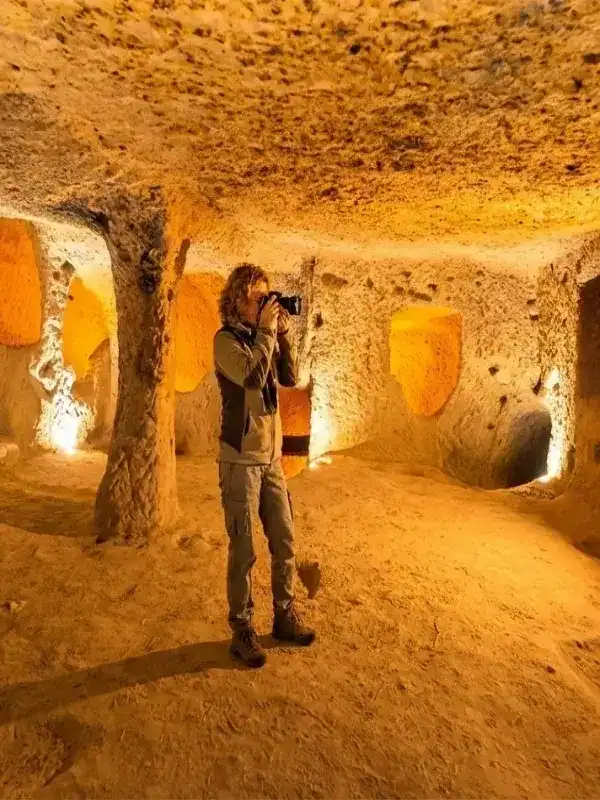 A female tourist with a camera exploring the intricately carved, warmly lit chambers of an ancient underground city, such as Derinkuyu or Kaymakli, in Cappadocia, Turkey.