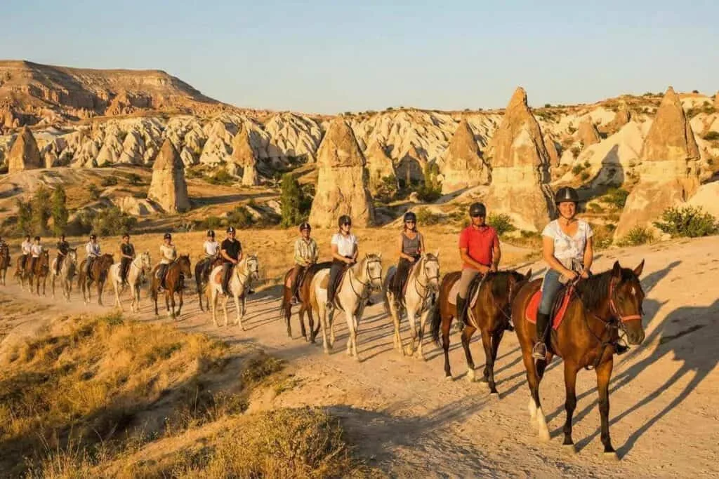 Tourists enjoying a sunset horseback riding tour in Cappadocia, Turkey, amidst scenic fairy chimney rock formations.