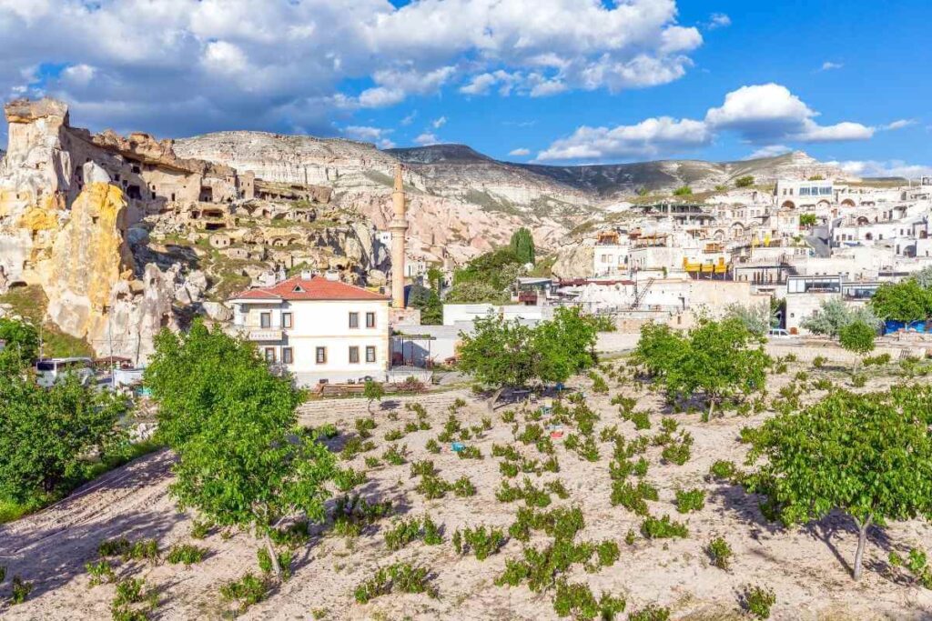 Scenic view of Çavuşin village in Cappadocia with cave dwellings, rock formations, and greenery under a bright blue sky.