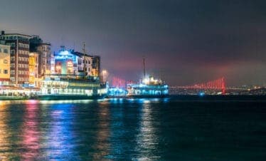 Colorful night view of Istanbul ferryboats docked by lively waterfront with illuminated Bosphorus Bridge in the background.