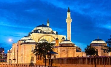 Illuminated Selimiye Mosque in Konya, Turkey at dusk with domes and minaret against a deep blue sky.