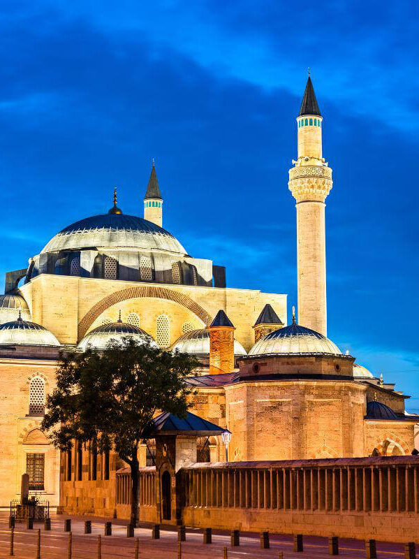 Illuminated Selimiye Mosque in Konya, Turkey at dusk with domes and minaret against a deep blue sky.