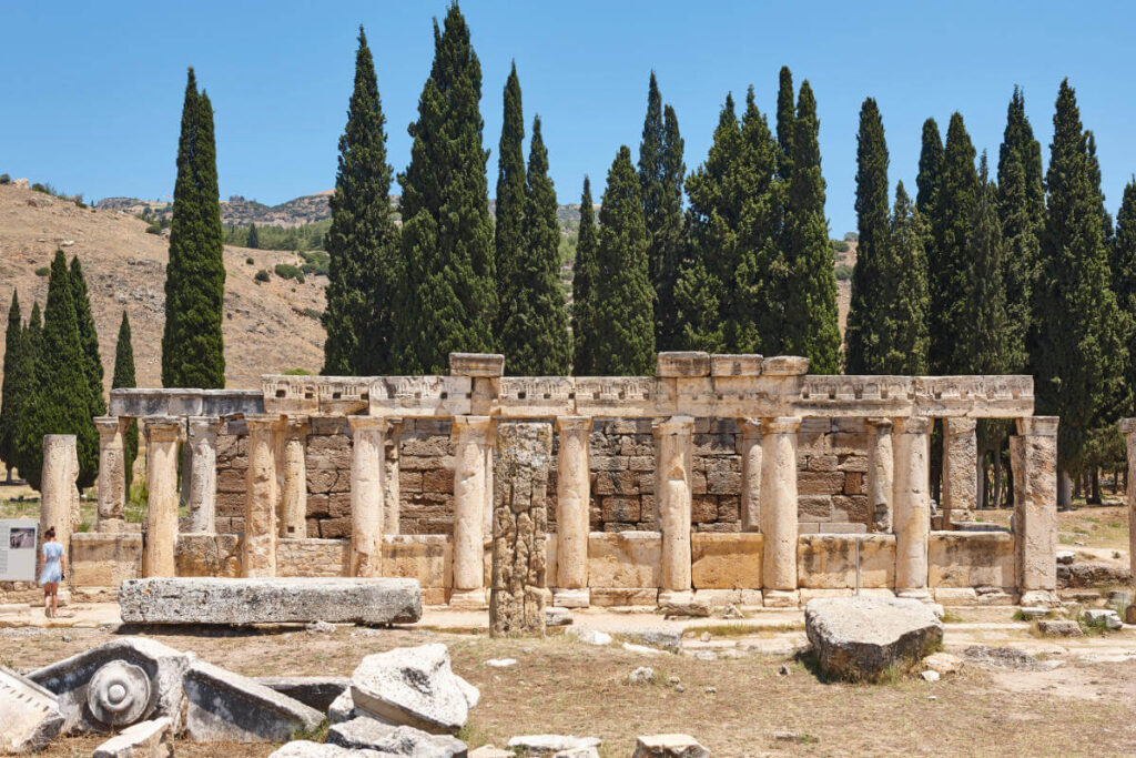 Stone-columned Roman latrine ruins in Hierapolis, Pamukkale, showing ancient public toilet structure amid cypress trees.