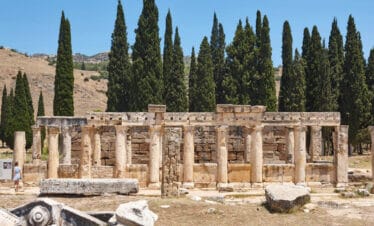 Stone-columned Roman latrine ruins in Hierapolis, Pamukkale, showing ancient public toilet structure amid cypress trees.