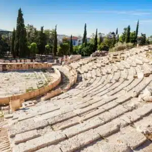 Ancient stone seating of the Theatre of Dionysus at the Acropolis, surrounded by trees and a clear sky, with distant mountains.