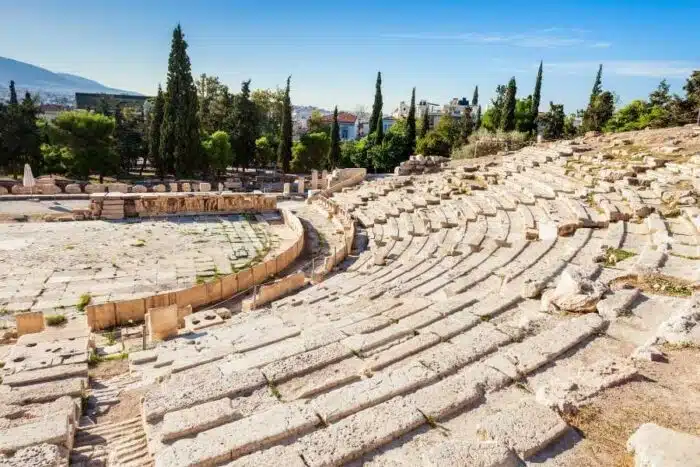 Ancient stone seating of the Theatre of Dionysus at the Acropolis, surrounded by trees and a clear sky, with distant mountains.