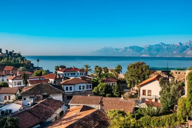 A scenic view of the historic Kaleici old town in Antalya, Turkey, featuring traditional white houses with red-tiled roofs overlooking the calm blue Mediterranean Sea and distant Taurus Mountains.