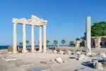 Ruins of the Temple of Apollo in Side, Turkey, with tall columns and a coastal view framed by palm trees and blue sky.