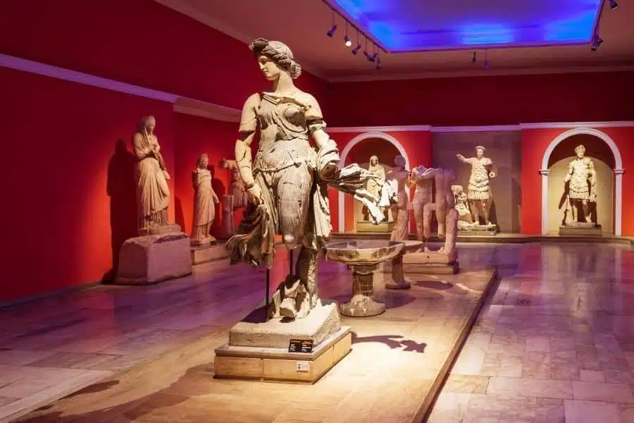 Ancient statues displayed in Antalya Museum, featuring a central armored figure surrounded by sculptures in a red-walled gallery.