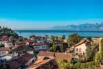 Rooftop view of Kaleiçi, Antalya, featuring traditional red-tiled houses, lush greenery, and the Mediterranean Sea with distant mountains.