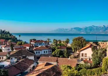 Rooftop view of Kaleiçi, Antalya, featuring traditional red-tiled houses, lush greenery, and the Mediterranean Sea with distant mountains.