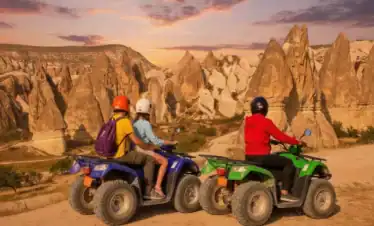 Two riders on a blue ATV and one rider on a green ATV pause on a dirt path to view the unique rock formations of Cappadocia, Turkey, under a dramatic orange and purple sunset sky.