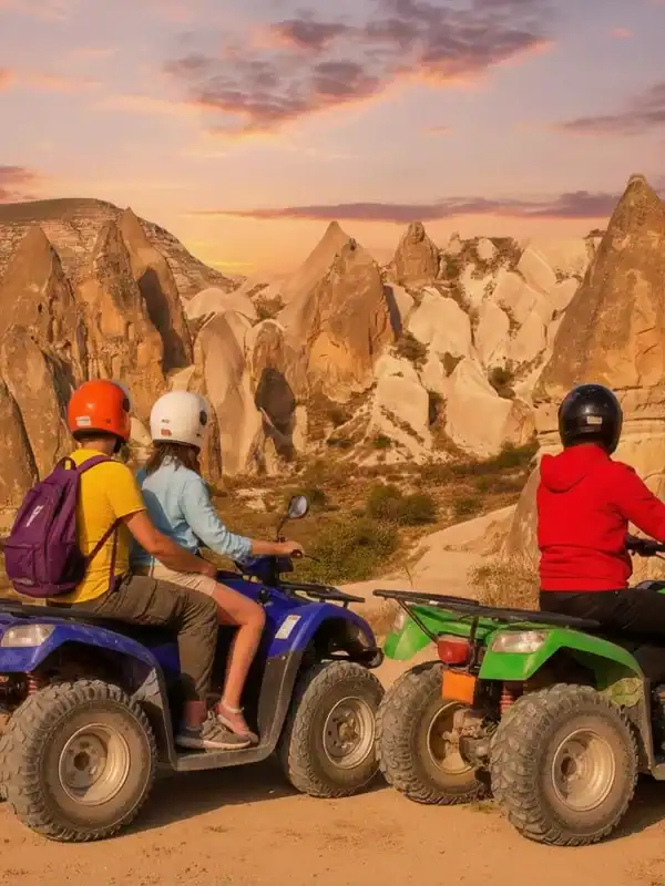 Two riders on a blue ATV and one rider on a green ATV pause on a dirt path to view the unique rock formations of Cappadocia, Turkey, under a dramatic orange and purple sunset sky.