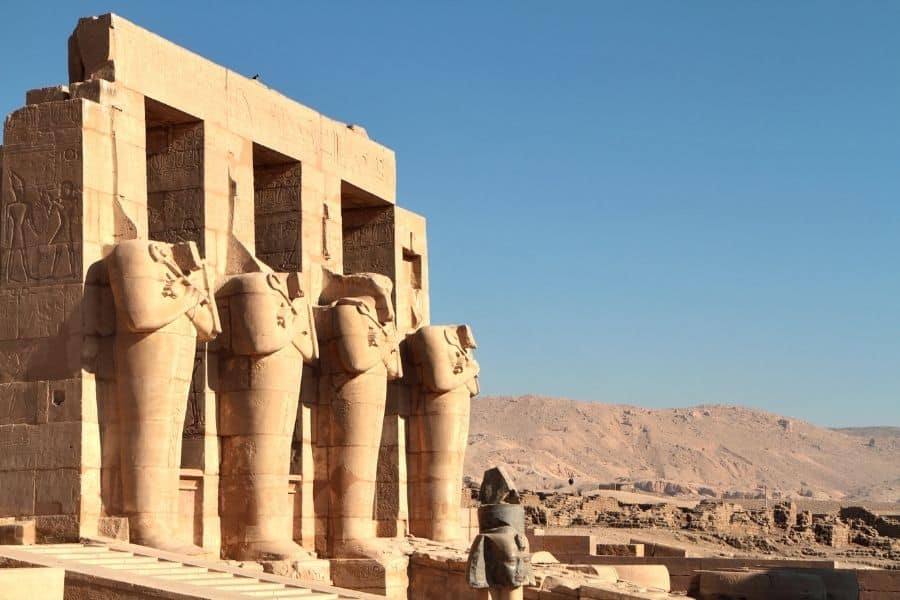 Stone statues of ancient Egyptian pharaohs at Theban Necropolis temple in Luxor, framed by desert landscape and clear sky.