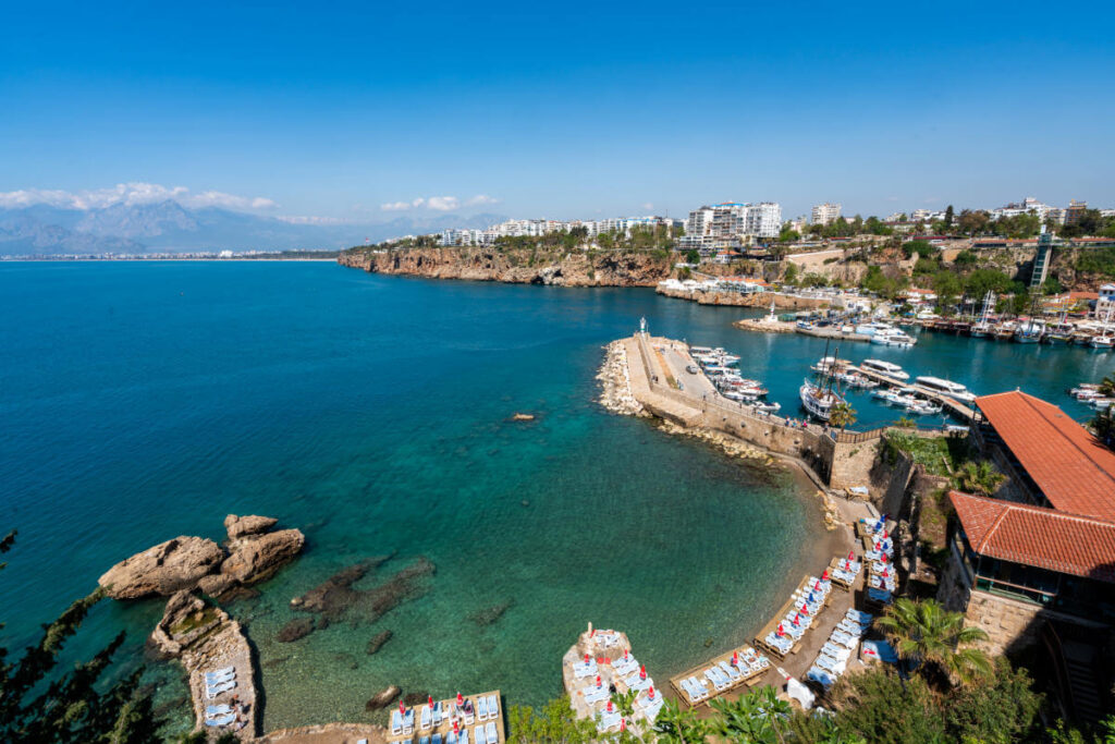 Stunning view of Antalya Old Harbor, Turkey, with clear turquoise waters, boats, cliffs, and beach loungers along the Mediterranean coast.