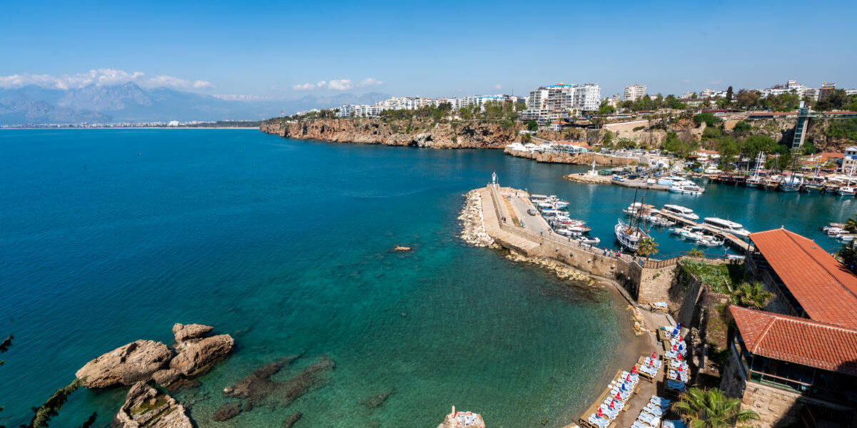 Stunning view of Antalya Old Harbor, Turkey, with clear turquoise waters, boats, cliffs, and beach loungers along the Mediterranean coast.