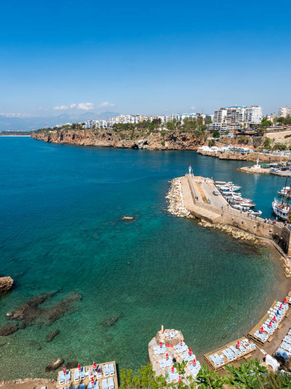Stunning view of Antalya Old Harbor, Turkey, with clear turquoise waters, boats, cliffs, and beach loungers along the Mediterranean coast.