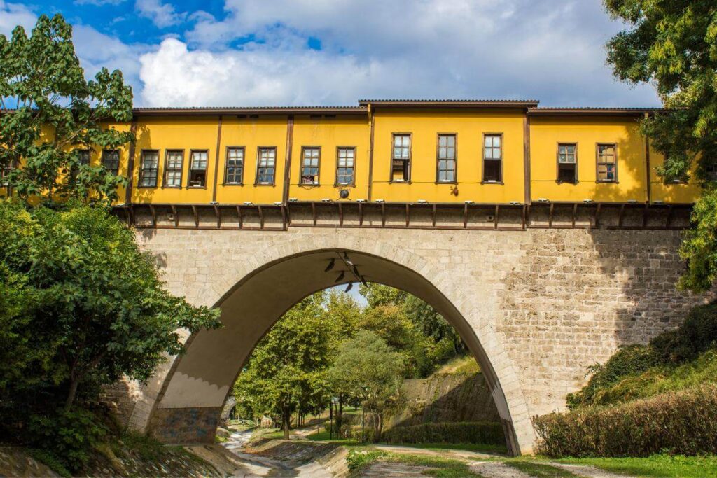 Yellow-painted Irgandı Bridge in Bursa with arched stone base and shops above, surrounded by green trees