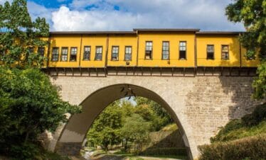 Yellow-painted Irgandı Bridge in Bursa with arched stone base and shops above, surrounded by green trees