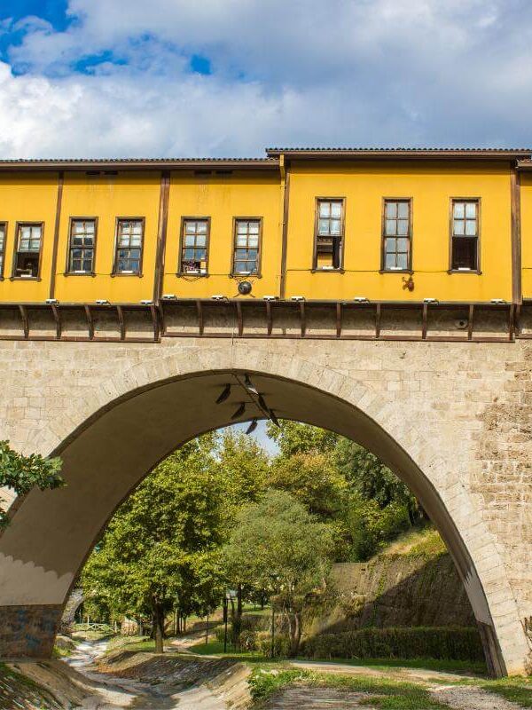Yellow-painted Irgandı Bridge in Bursa with arched stone base and shops above, surrounded by green trees