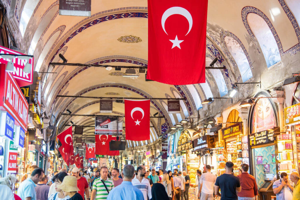 Shoppers browse vibrant stalls under arched ceilings with Turkish flags in Istanbul’s historic Grand Bazaar.