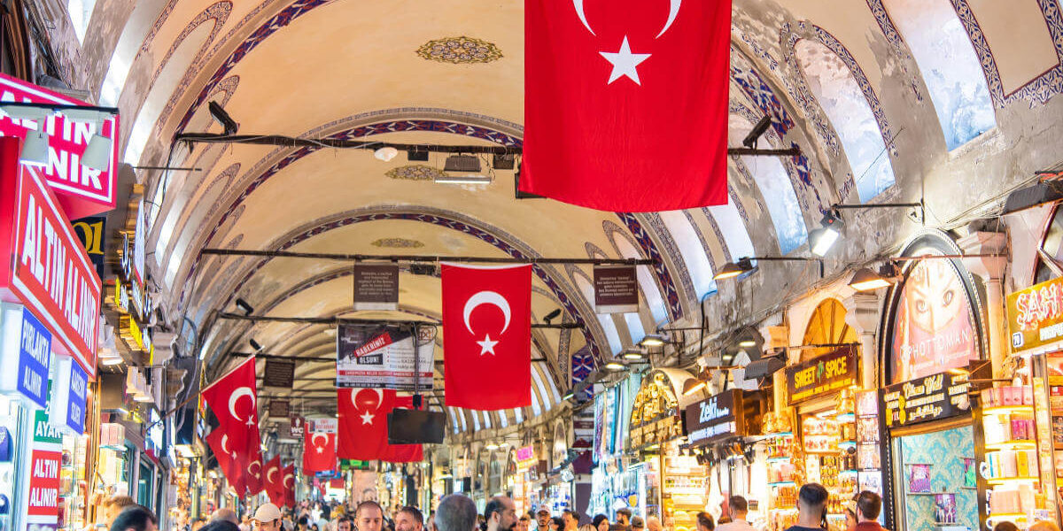 Shoppers browse vibrant stalls under arched ceilings with Turkish flags in Istanbul’s historic Grand Bazaar.