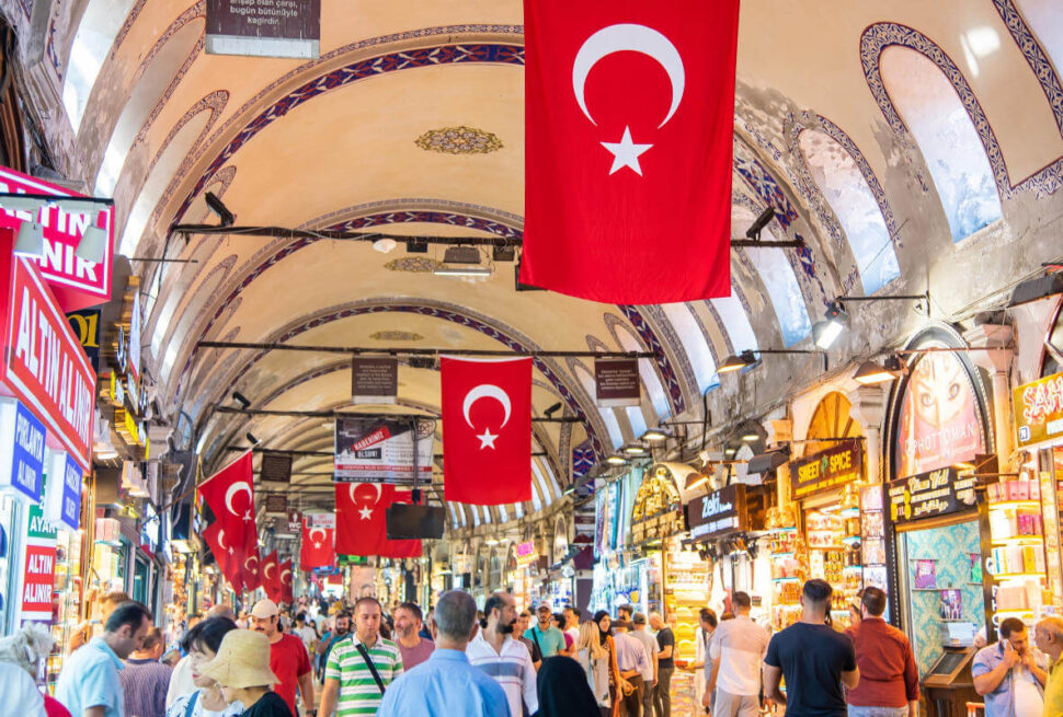 Shoppers browse vibrant stalls under arched ceilings with Turkish flags in Istanbul’s historic Grand Bazaar.