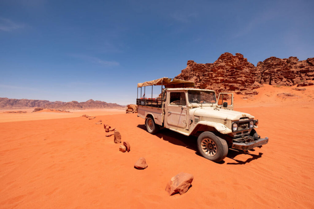 Vintage white jeep parked on red sand in Wadi Rum desert with rocky cliffs and clear blue sky in the background.