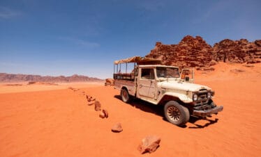 Vintage white jeep parked on red sand in Wadi Rum desert with rocky cliffs and clear blue sky in the background.