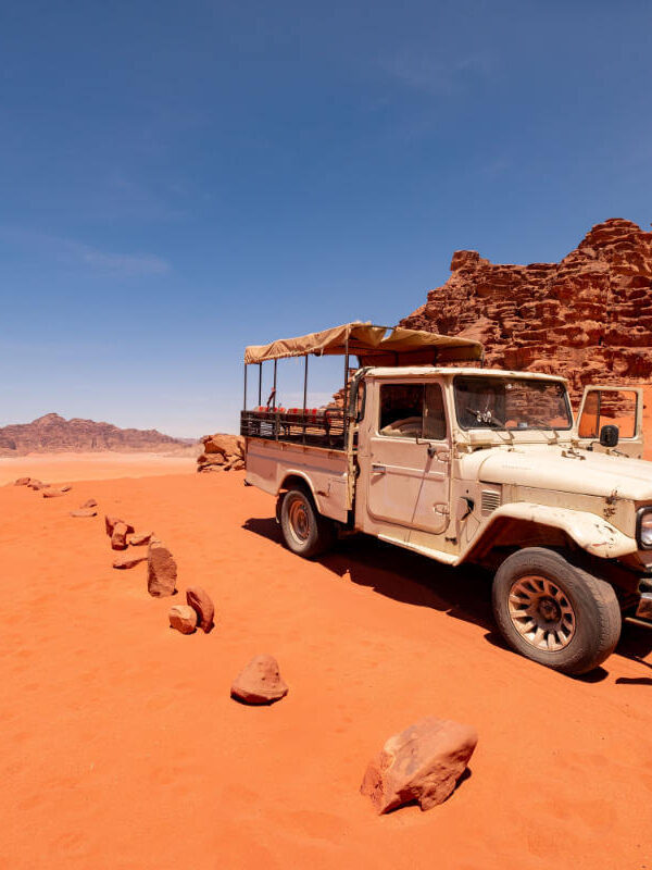 Vintage white jeep parked on red sand in Wadi Rum desert with rocky cliffs and clear blue sky in the background.