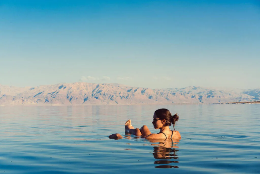 Woman floating effortlessly in the Dead Sea with mountain backdrop and clear blue sky.