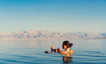 Woman floating effortlessly in the Dead Sea with mountain backdrop and clear blue sky.