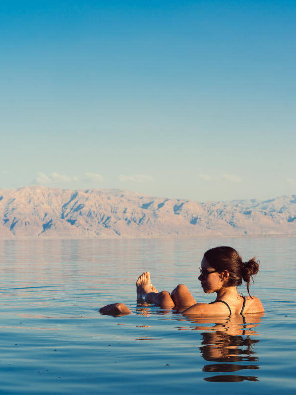 Woman floating effortlessly in the Dead Sea with mountain backdrop and clear blue sky.