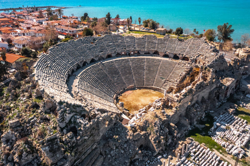 Aerial view of the Ancient Theater of Side in Antalya, Turkey, with stone seating and coastal town in the background.