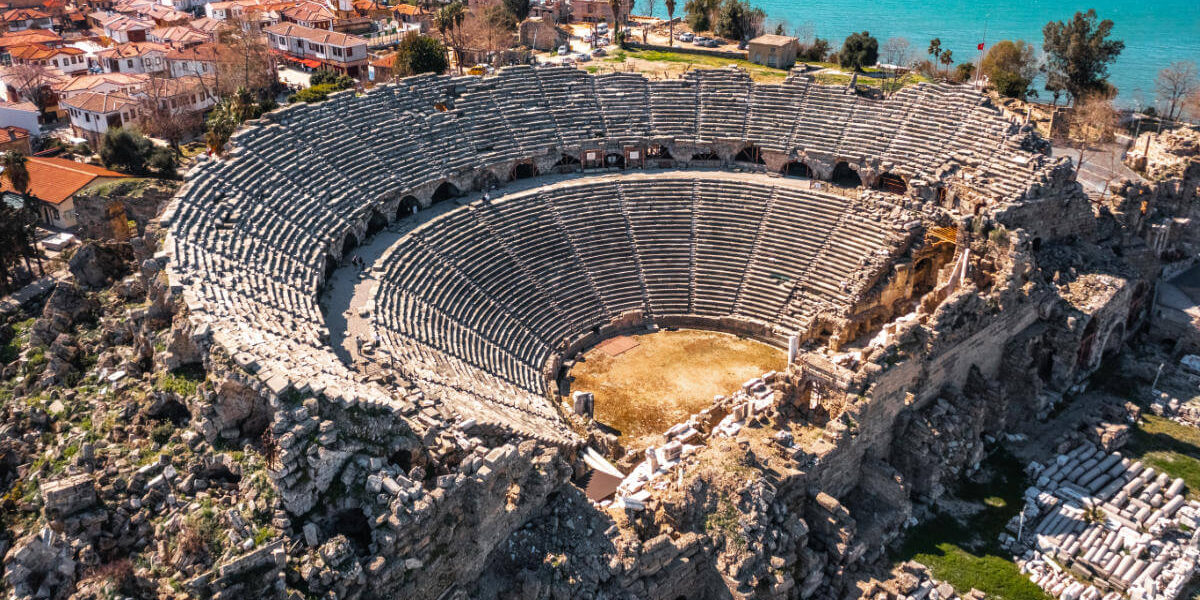 Aerial view of the Ancient Theater of Side in Antalya, Turkey, with stone seating and coastal town in the background.