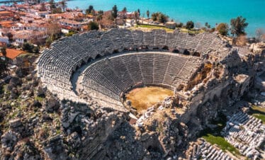 Aerial view of the Ancient Theater of Side in Antalya, Turkey, with stone seating and coastal town in the background.